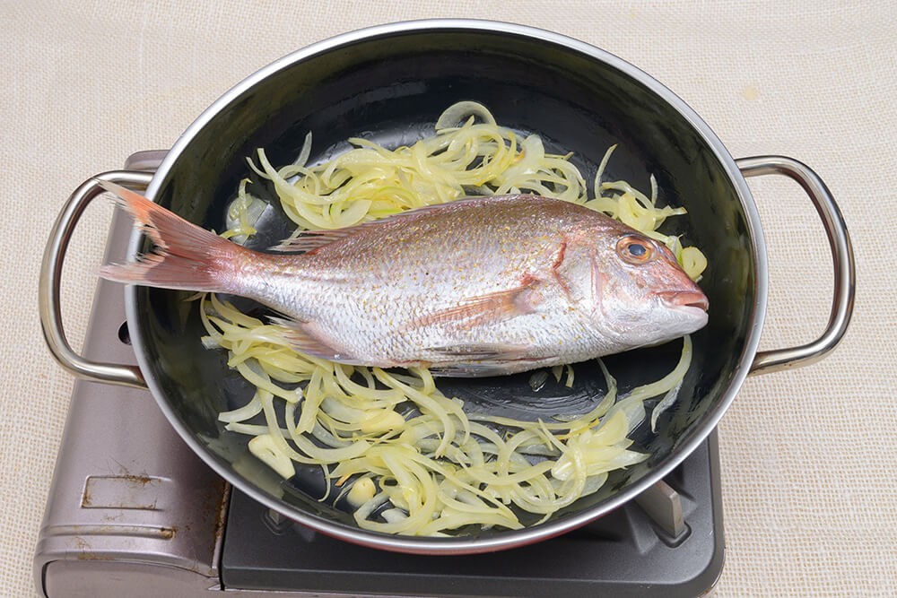 油がまわったら具材を両端によせ、盛り付ける側を下にして鯛を焼く。焼き色が付いたらひっくり返し、鯛のまわりに3の具材とあさりを入れ、白ワイン、水を加えてフタをし、中火にかける。